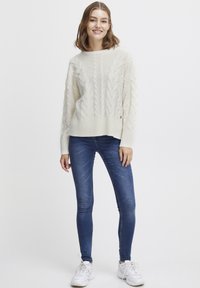 Pulover de culoare crem, tricotat cu model cable-knit, cu tivuri și manșete cu garnitură, purtat cu blugi skinny albaștri și adidași albi, stând în fața unui fundal simplu.