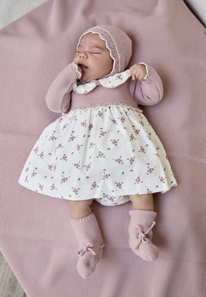 Bebé durmiendo con un gorro de punto rosa, suéter, botines y un vestido blanco con flores, acostado sobre una manta texturizada rosa.
