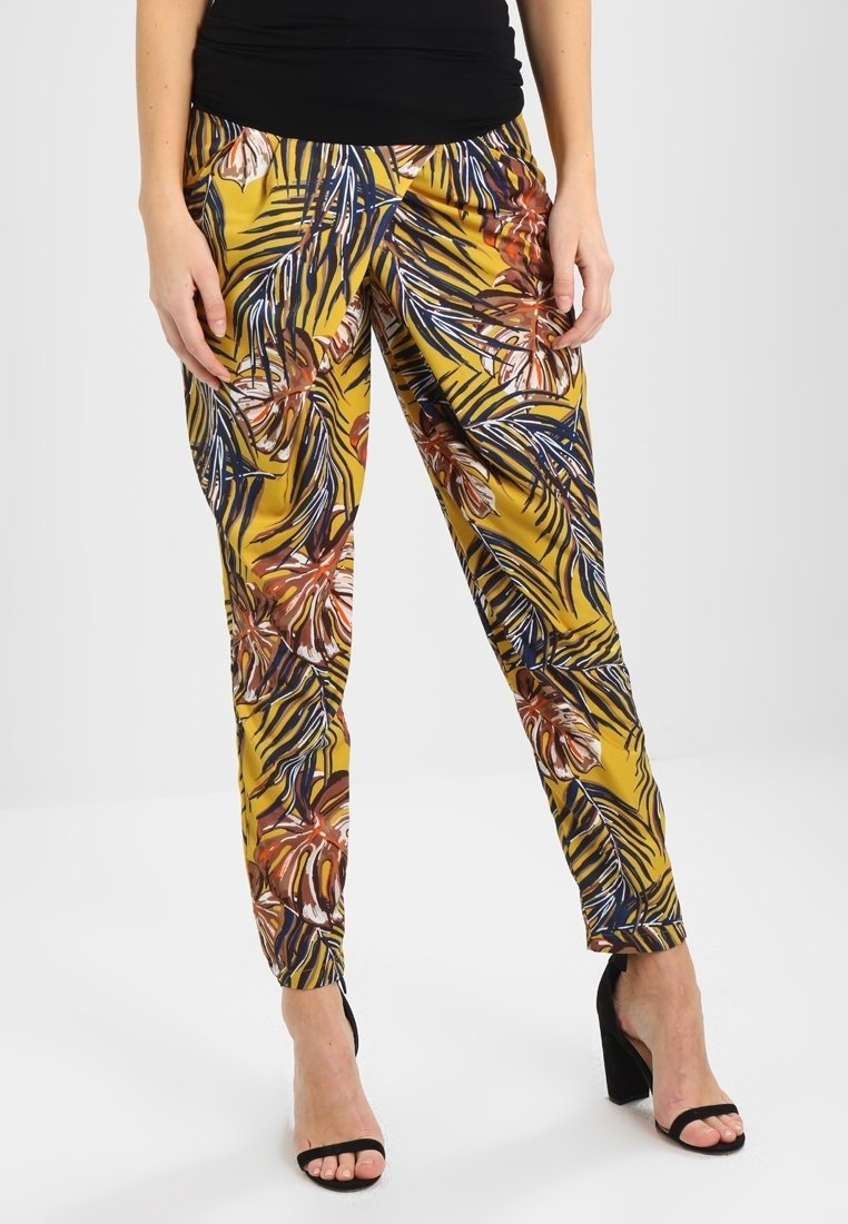 Femme portant un pantalon jaune évasé avec un imprimé feuilles tropicales multicolore et des sandales à talons noirs, debout devant un fond uni.