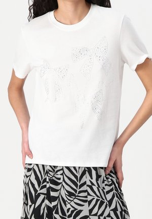 Camicia bianca a maniche corte con tre motivi a fiocco di strass abbinata a una gonna con motivo di foglie in bianco e nero su una persona.