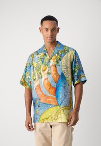 Joven con una camisa de botones de manga corta azul con un estampado colorido de manos y naturaleza, combinada con pantalones beiges, de pie contra un fondo liso.