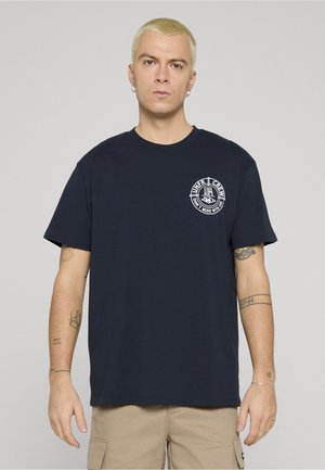 Lühikeste blondide juustega mees kannab musta graafilist T-särki ja beeže pükse, seistes tavalise heleroheka tausta ees.