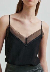 Nainen, jolla on päällään musta hihaton paita, jossa on ohuet hihnat ja läpikuultava V-muotoinen kaula-aukko, taustanaan yksinkertainen vaalean värinen tausta.