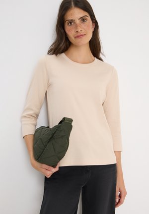Haut beige à manches longues en maille côtelée avec un col rond, porté avec un pantalon noir ; tenant un petit sac à épaule en quilté vert.