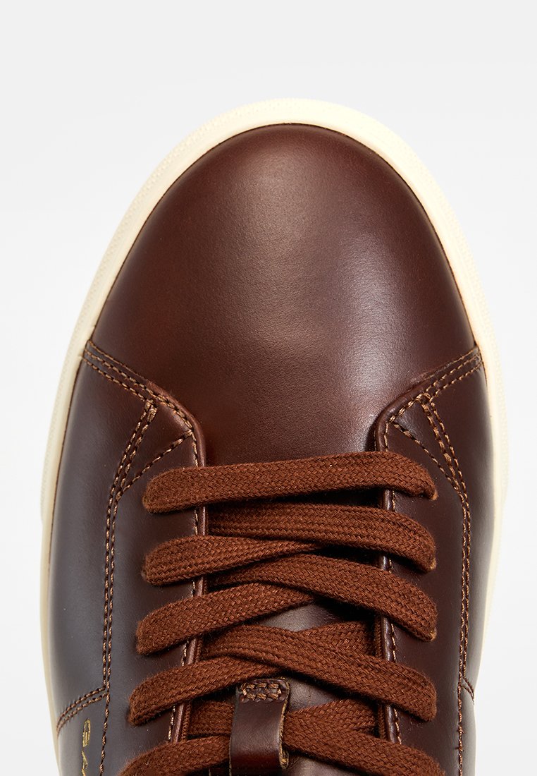 Brun lädersneaker med en slät yta, krämfärgad gummisula och bruna snören. Har enkel sömnad och en rund tådesign.