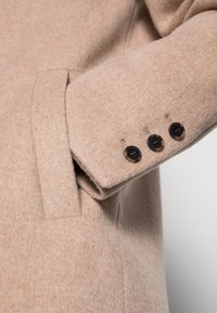 Manche d'un manteau en laine beige avec trois boutons foncés et une poche fendu verticale sur le panneau latéral.