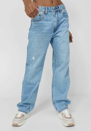 Personne portant un jean bleu clair coupe décontractée avec un léger détail usé et des baskets blanches, debout devant un fond uni.