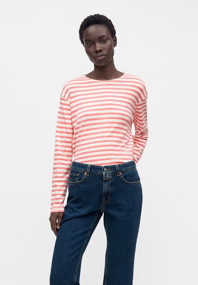Femme portant une chemise à manches longues rose et blanche rayée et un jean taille haute bleu foncé, se tenant devant un fond blanc uni.