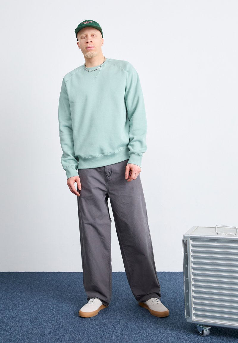 Sweatshirt vert clair avec un col rond, pantalon large gris et baskets beiges. Valise à roulettes argentée à côté. Fond uni.