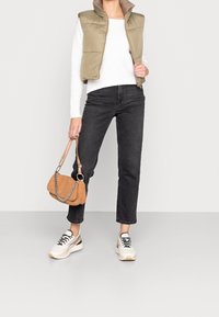 Zelená olivově polstrovaná vesta přes bílý žebrovaný svetr, doplněná černými zkrácenými džíny a bílými teniskami. Drží hnědou kordurovou kabelku.