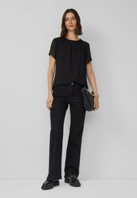 Vrouw met een zwarte blouse met korte mouwen en zwarte broek, die een zwarte handtas met een gouden kettingriem vasthoudt, staand voor een lichtgrijze achtergrond.