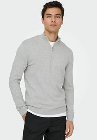 Pull à col zippé gris clair avec des poignets et un ourlet côtelés, fabriqué en matière douce, porté sur une chemise blanche, associé à un pantalon noir.