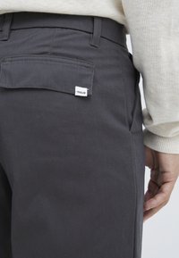 Donkergrijze broek van stevig materiaal, met een platte achterzak met een klein label met de tekst "!SOLID." Eenvoudig en functioneel ontwerp.