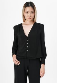 Jeune femme avec des cheveux raides noirs et blonds portant un blouse boutonnée noire à col en V et un pantalon noir sur un fond blanc.