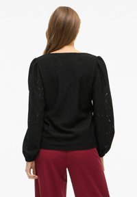Blusa nera texturizzata con maniche lunghe a palloncino, in tessuto a motivo diamante, con scollatura quadrata e vestibilità rilassata. Abbinata a pantaloni bordeaux.