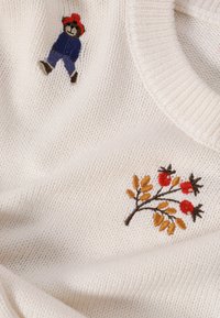 Biały dziergany sweter z haftowanym misiem w niebieskim i czerwonym stroju, obok kwiatowego wzoru z czerwonymi jagodami i brązowymi liśćmi.