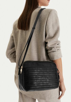 Donna che indossa un blazer beige con una borsa a tracolla in pelle nera con strisce orizzontali in rilievo e dettagli in oro sulla cerniera.