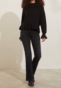 Un pull en tricot noir avec un ourlet froncé et des manches en cloche, associé à un jean foncé ajusté et des bottines, sur un fond neutre.