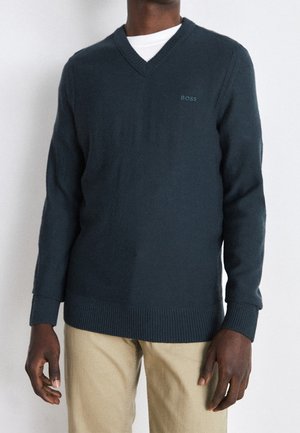 Man wearing dark blue V-neck sweater with "BOSS" logo over white undershirt and beige pants against plain background.