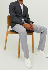 Blazer a quadri scuri con rever, abbinato a pantaloni della tuta grigi chiari e sneakers bianche. Modello seduto su una sedia di legno.