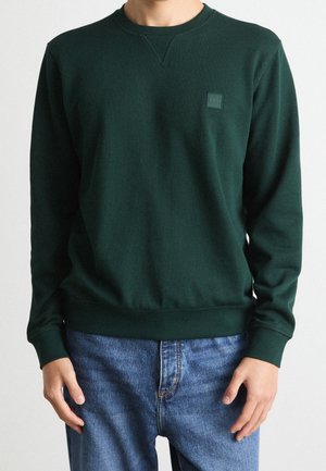 Personne portant un sweat-shirt col rond vert foncé avec un petit logo « BOSS » sur la poitrine et un jean bleu, debout devant un fond uni.