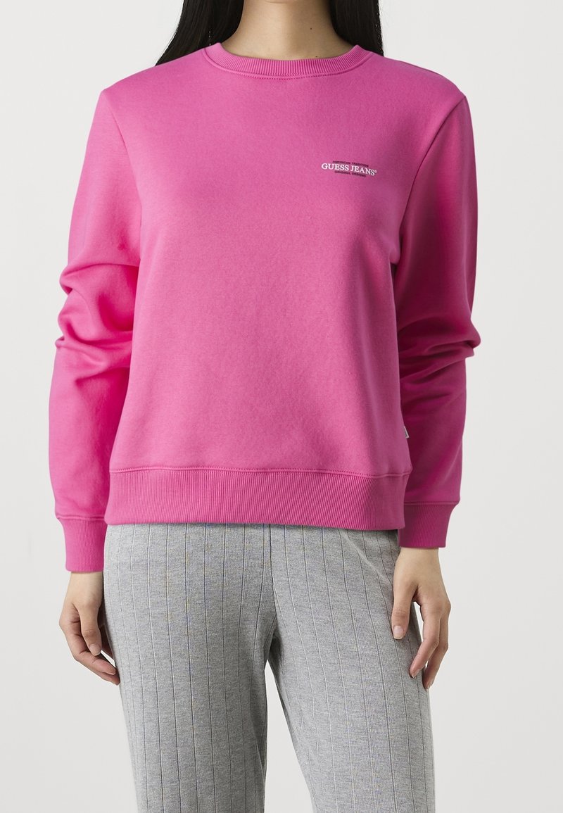 Femme portant un sweat-shirt Guess Jeans rose vif avec poignets, ourlet et col côtelés, associé à un pantalon gris à fines rayures.