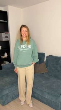 Πράσινο φούτερ με κουκούλα με το λογότυπο "Ripcurl Surf Sports", κρεμ πλεκτό παντελόνι, λευκά αθλητικά παπούτσια, που στέκεται σε ανοιχτόχρωμο ξύλινο πάτωμα δίπλα σε υφασμάτινο μπλε καναπέ.