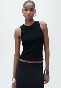 Black ribbed tank top with a round neckline and sleeveless design. Worn with a black skirt and metallic bracelets on both wrists.