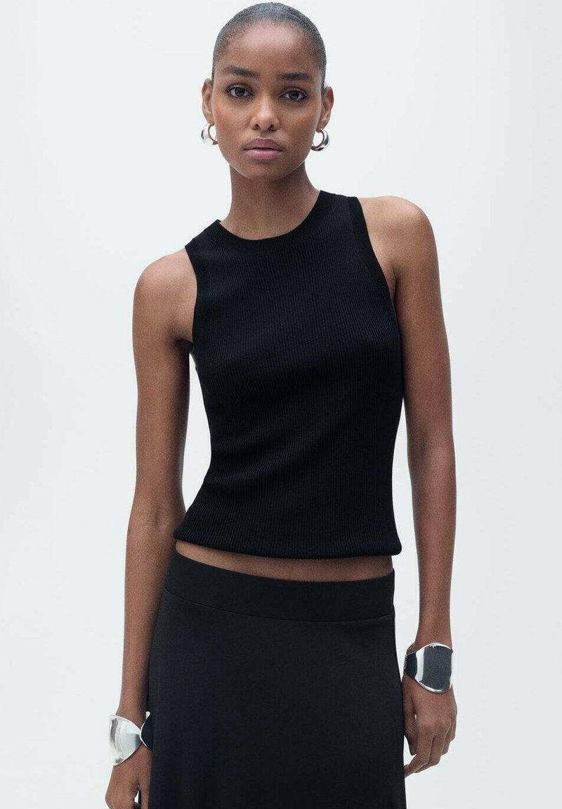 Black ribbed tank top with a round neckline and sleeveless design. Worn with a black skirt and metallic bracelets on both wrists.