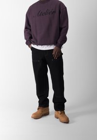 Lilla sweatshirt med teksten "Atelier", parret med sorte bukser med synlig syning og brune arbejdssko med snørebånd.