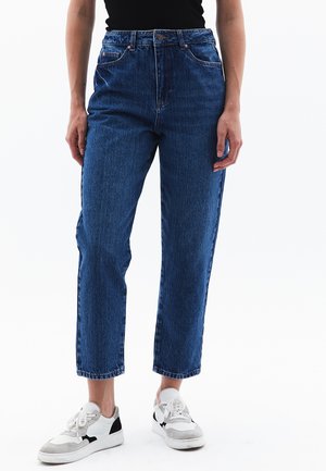 Jean en denim bleu taille haute avec une coupe droite, présentant un design cinq poches et des coutures contrastantes, associés à des baskets blanches et grises.