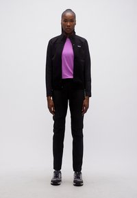 Γυναίκα που στέκεται με προσανατολισμό προς τα εμπρός, φορώντας μαύρο μπουφάν, μοβ πουκάμισο, μαύρο παντελόνι και γκρι αθλητικά παπούτσια, με φόντο απλό λευκό.