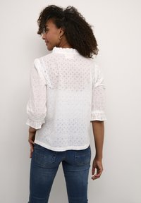 Blusa bianca in cotone con motivi ad occhiello, colletto con volant e maniche a sbuffo. Abbinata a jeans blu aderenti, che mostrano uno stile casual.