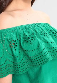 Zelený top bez ramínek s jemně vyšívanou krajkovou detailem a nařaseným výstřihem. Tkanina vypadá lehce a prodyšně.