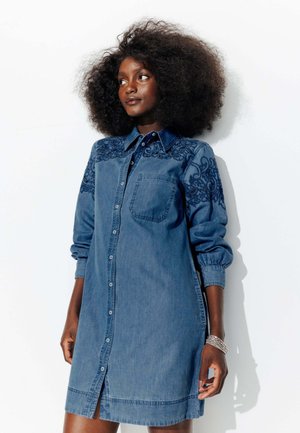Robe chemise en denim avec fermeture à boutons, manches longues et poche poitrine. Présente une broderie bleue complexe sur les épaules et les poignets.