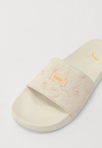 Sandale à glissière avec un dessus en daim rose pâle présentant des motifs floraux en relief, un logo orange et une semelle en caoutchouc beige.