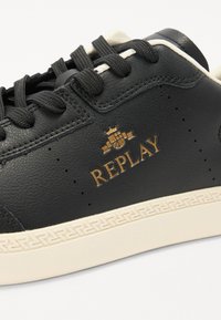 Black leather sneaker with cream sole, featuring black laces, perforated detailing, and gold "REPLAY" logo. Textured surface, rounded shape.