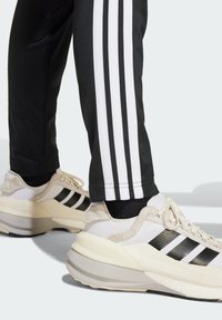 Baskets blanches avec des rayures noires, en mesh texturé et accents en daim, associées à un pantalon noir avec des rayures blanches sur les côtés.