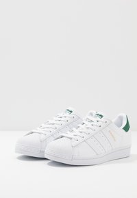 Vita lädersneakers med grön hälaccent, texturerad tåkupp, tre distinkta sidostripes och "SUPERSTAR"-märkning i guld.