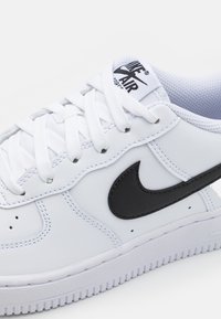 Baskets blanches Nike Air Force 1 avec logo swoosh noir, lacets blancs et semelle texturée sur un fond clair uni.