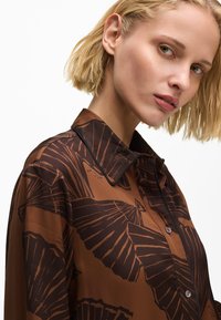 Rjava s posipom s cvetličnim vzorcem, z gumbi spredaj in detajlom ovratnika. Narejena iz lahkega blaga z gladko teksturo.