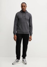 Felpa grigia con cappuccio e tasca frontale, logo nero, abbinata a pantaloni neri a sigaretta e scarpe sportive grigie con suola bianca.