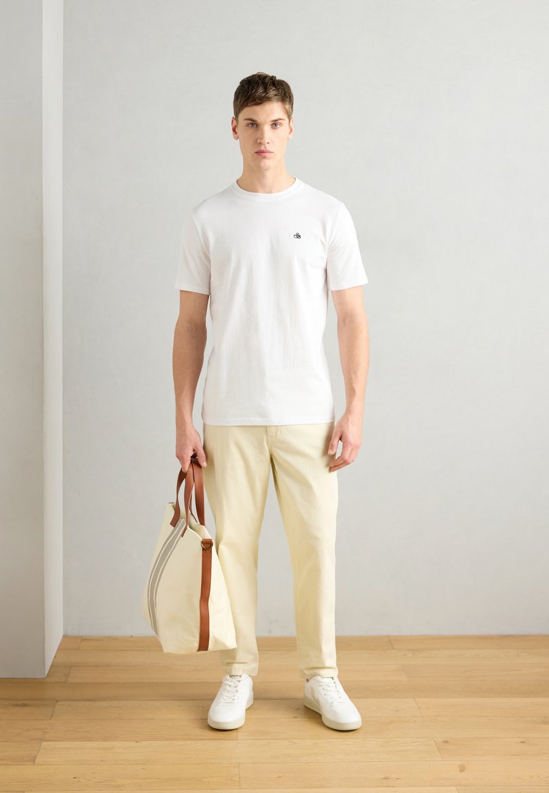 Hvid bomuld T-shirt med et lille logo, lysebeige bukser og hvide sneakers. Holder en cremefarvet taske med brune læderhåndtag.