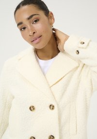 Manteau double boutonnage crème en tissu texturé, avec de grands boutons dorés, des revers crantés et des poches latérales. Apparence douce et confortable.