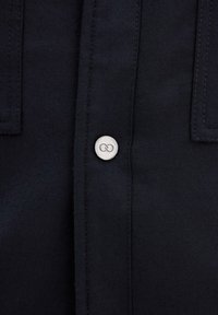 Camicia nera in tessuto morbido, con un bottone d'argento a forma di simbolo dell'infinito sulla parte superiore. Include due tasche sul petto.