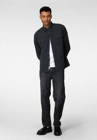 Svarte skjorte med knappestolpe og to brystlommer, brukt over en hvit skjorte, kombinert med mørkeblå jeans og svarte sko. Glatt stofftekstur.