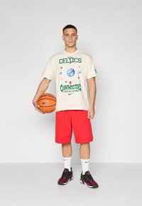 Kremfarget t-skjorte med Boston Celtics-logo og grafikk, kombinert med røde basketballshorts. Personen holder en basketball og har på seg svarte og røde sneakers.