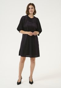 Mörklila A-linjeformad klänning med vertikala veck, rund halsringning och korta, puffiga ärmar. Bärs med svarta texturerade högklackade skor.