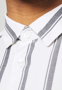 Camicia bianca con bottoni, caratterizzata da strisce verticali grigie, colletto standard e una texture liscia, con una chiusura a bottone singolo al collo.