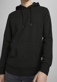 Czarna bluza z miękkiego materiału, wyposażona w kieszeń kangurka, regulowane sznurki oraz ściągacze przy dole i rękawach.